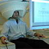 Photo of a person sitting in a chair with a large cylindrical apparatus around their head, looking at a computer screen and holding a response device.