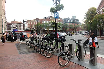 Street scene in a busy urban square, with parked Hubway bicycles, cars on street, pedestrians on sidewalk and crossing the street, and entrance to a subway stop in the distance.