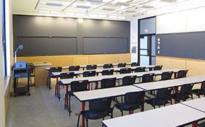 Five rows of narrow white tables and black chairs facing the front of the classroom. Sliding chalkboards at the front and side of the classroom.
