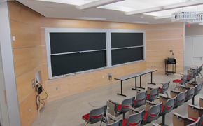 Tiered lecture hall with tablet arm desks, and chalkboards and projector at the front of the room.