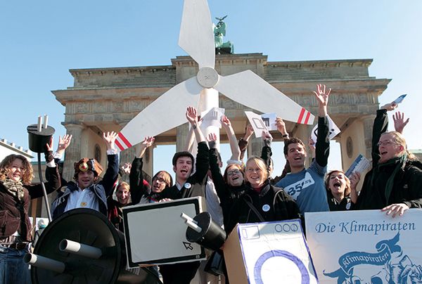 Ten people standing in front of the Brandenburg Gate. They are raising their hands in the air holding props and standing in front of a model of a windmill.