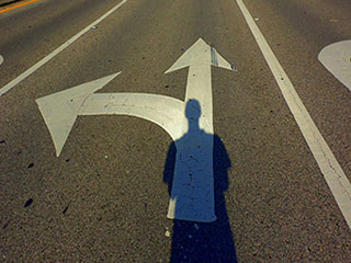 A man's shadow falls on two arrows painted on a road—one arrow points left and the other points straight ahead.