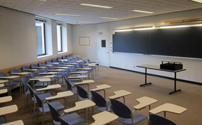 Classroom with three curved rows of desks facing a small table in front of a chalkboard; a lectern is placed on top of the table.