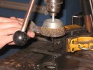 A drill press with a rotating wire brush scrubs rust off a bottle opener.