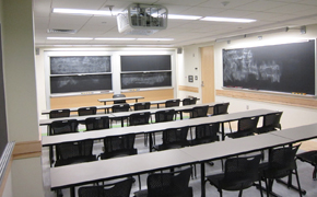 Classroom with three chalkboards and four long tables with 19 modern black chairs visible.