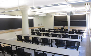 A classroom with five long tables spanning the classroom, chairs behind each table, and two sliding chalkboards at the front.