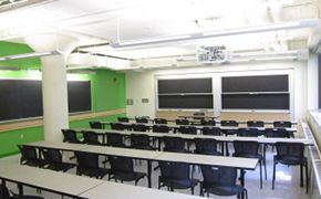 Four rows of grey tables with black chairs. Sliding blackboards at the front and along the left hand side of the room. The wall to the left is painted green.