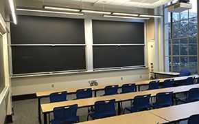 A classroom with rows of tables and chairs facing four large blackboards.