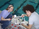 Two students posing with a cube-framed robot at the edge of the pool.