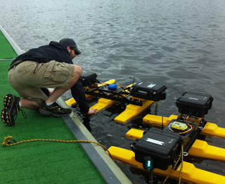 A man crouched on a river dock, reaching down to two small floating “robots” that consist of several boxes of electronics supported on pontoons.