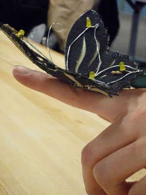 Photo of a fabric butterfly with several LEDs on its wings, perched on a persons hand.