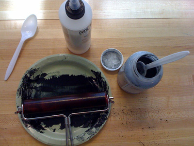 Photo of black paint on a plate, with roller, and a couple of containers. 