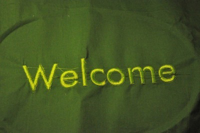 Photo of fabric embroidered with the word Welcome in a Helvetica-style font. 