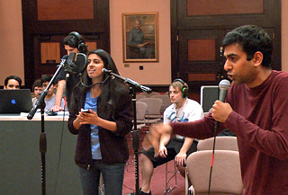 Photo of two singers on microphones, with a recording engineer standing in the background monitoring the session on a laptop.
