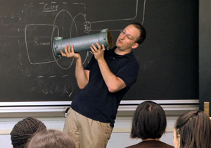 Dr. Short stands in front of the blackboard holding a metal tube about six inches thick.