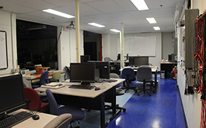 Computer monitors on grey tables. Lab equipment also on some of the tables. Moveable chairs. White boards on the walls. Red and black cables hanging on the right wall of classroom.