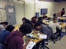 Photo of a classroom with 3 rectangular tables, 11 students seated around them, soldering and assembling electronics.