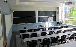 A classroom with sliding chalkboards at the front, and around 40 chairs arranged in arrows at long tables.