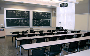 Photo of a classroom with long tables arranged with chairs so that they face the four chalkboard panels in the front of the room.