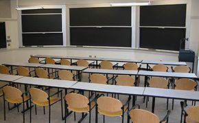 Three rows of long grey tables with wooden chairs facing chalkboards.