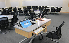 View of classroom from front shows computer, overhead, 8-top round student tables, and whiteboards around the entire room.