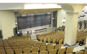 View of a lecture hall from the rear of the classroom. Chairs are affixed to the floor and are tiered. There are two aisles between three sections of seating. Blackboards at the front of the classroom. 