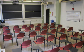In this medium-sized classroom, six rows of seats are visible. Each row has six to seven chairs with attached desks. At the front, there is a small table and two columns of sliding chalkboards.
