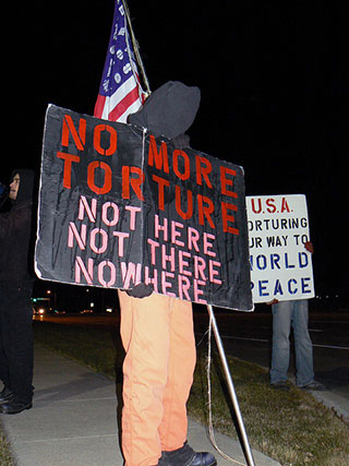 A protester stands with a black hood over his face while holding a sign that reads “No more torture, not here, not there, nowhere.”