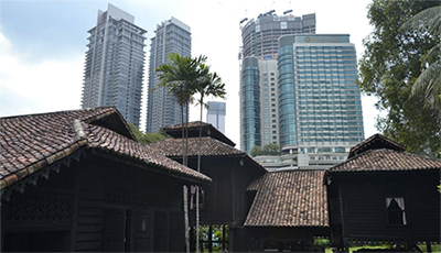 A traditional Malaysian house is in the foreground, with skyscrapers in the background. 
