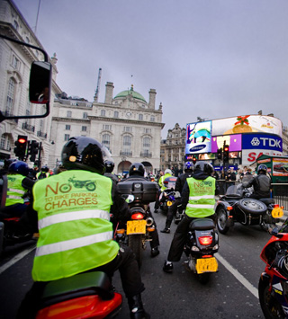Photo of a large group of motorcyclists riding down a city street, some wearing vests with protest message 'No to bike parking charges!'