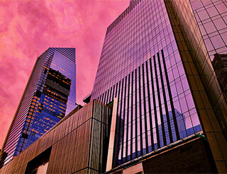 Two glass buildings rise into the sky. They glow pink as they reflect the sunset. 
