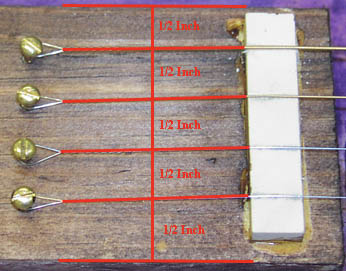 Four guitar strings stretched over tailpiece nut blank, with measurements.