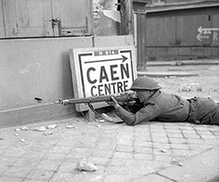A European soldier lies on his stomach aiming a rifle. There is rubble around him and a sign pointing towards Caen Centre.