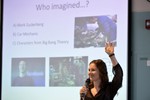A woman holding a microphone is giving a presentation and raising her hand. The slide presentation in the background says "Who imagined...? Mark Zuckerberg, Car Mechanic, Characters from Big Bang Theory."