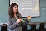 A woman holds a microphone and gives a lecture in front of a slide show. She is holding a rough prototype of a prosthetic foot.