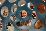 A girl is holding a 3D printed plastic object with holes in it up to the camera.
