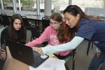 Girls sit around a laptop, and a volunteer is standing and helping at the computer.