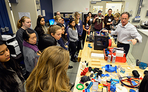 Twenty girls stand near a wooden table. Several items printed with a 3D printer lay on the table. A scientist stands behind the table and gestures toward the items.