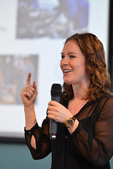 A photo of Kristen Railey giving a presentation, speaking into a microphone with a projected slide in the background.