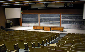 Large lecture hall as seen from the back of the room; approximately 230 chairs with writing tablets face a row of 3 tables at the front of the room. Three large sliding blackboards are behind the row of tables. A large white screen is to the side of the blackboards.